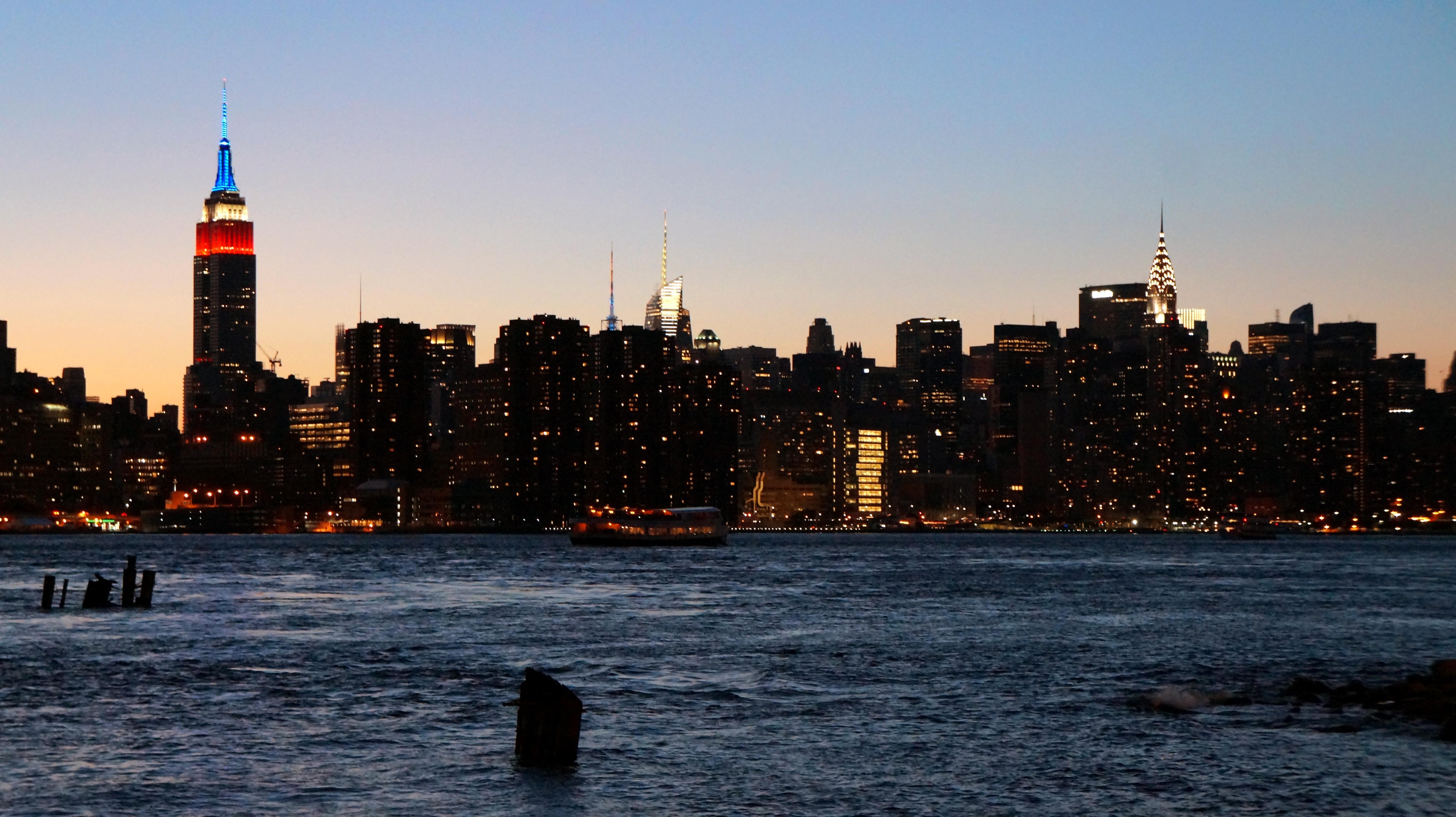 The New York City skyline at dusk featuring the Empire State Building and Chrysler Building illuminated above dark water.