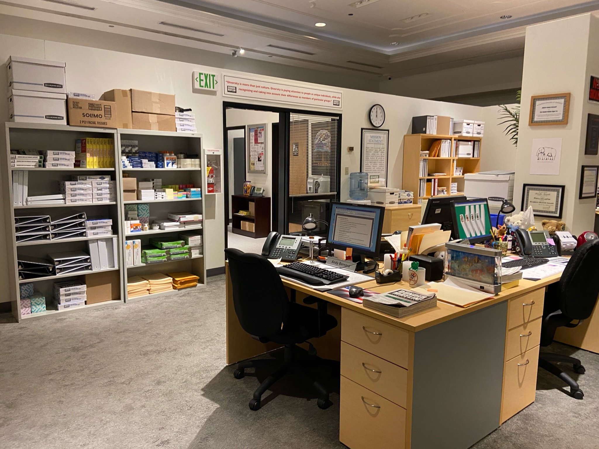 A full-scale office set recreation featuring desks with computer monitors and supplies in front of shelving units and an office doorway.
