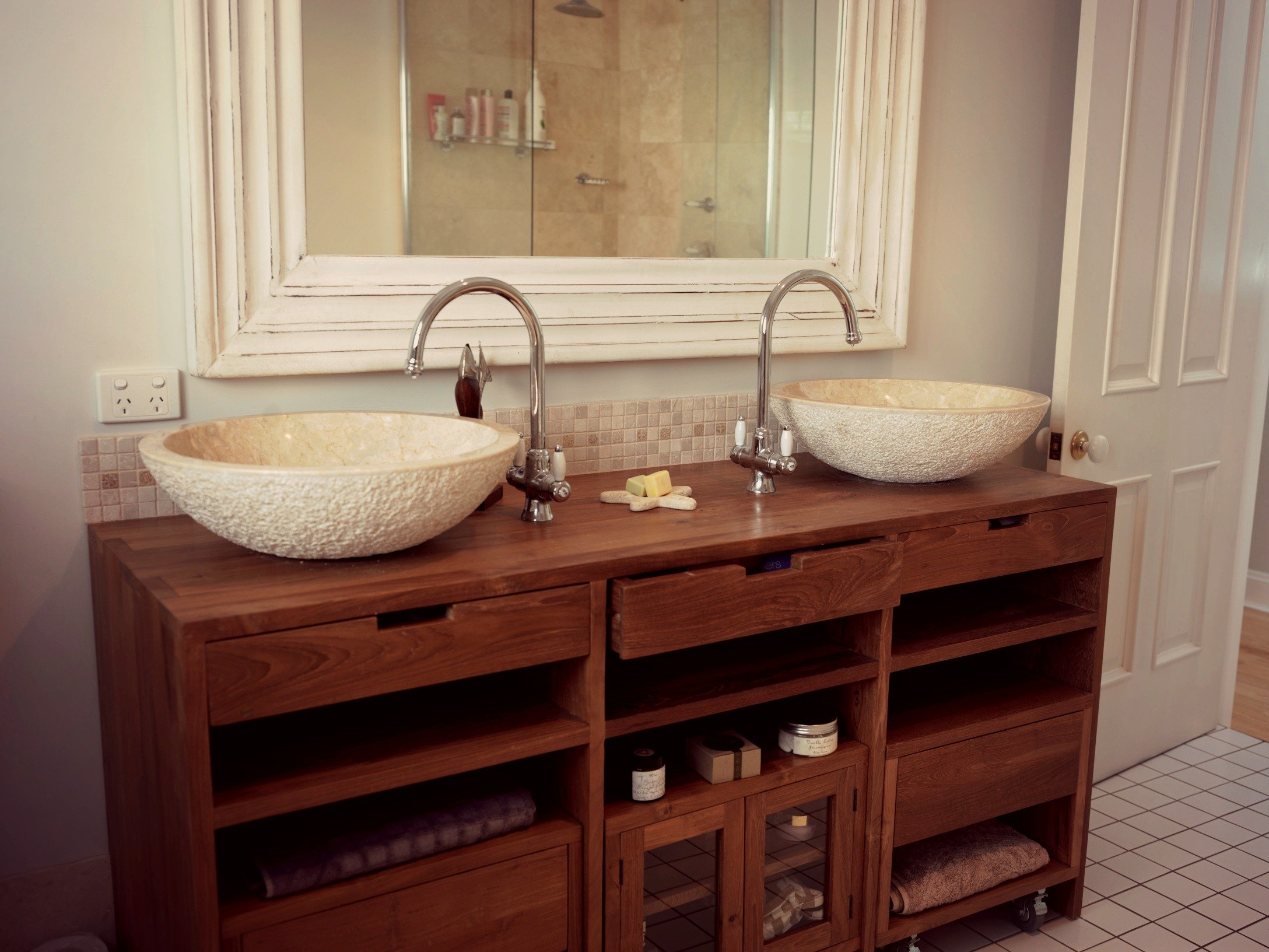 A rustic double vanity with stone vessel sinks and chrome faucets in a bathroom with white tiled flooring.