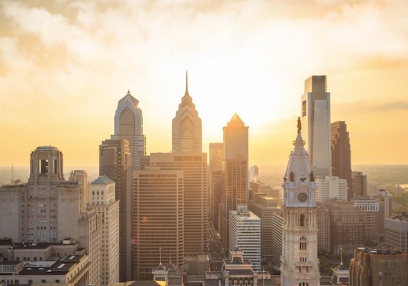 The Philadelphia skyline at sunrise with golden light illuminating One Liberty Place, the Comcast Center, and the City Hall clock tower.