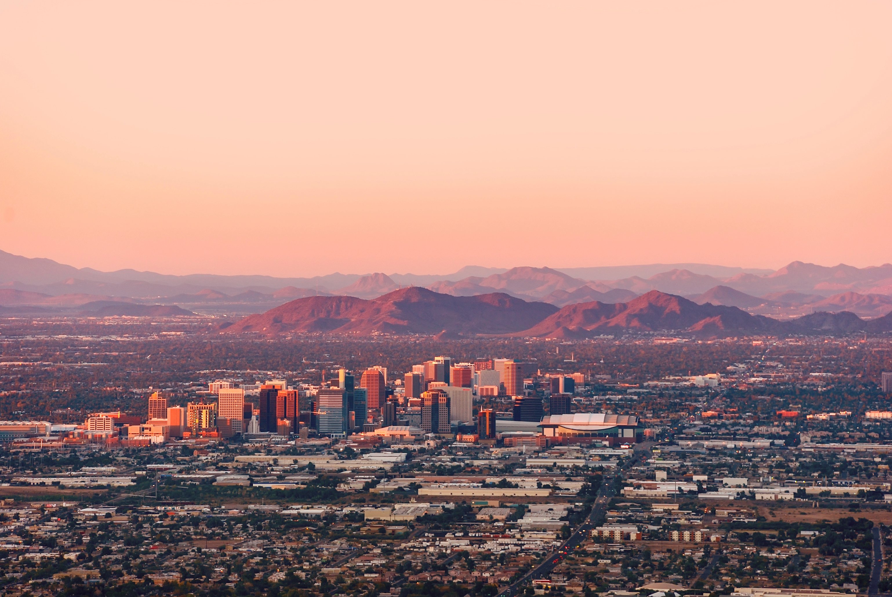 Aerial view of the Phoenix downtown skyline and urban valley illuminated by sunset light against a backdrop of desert mountains.