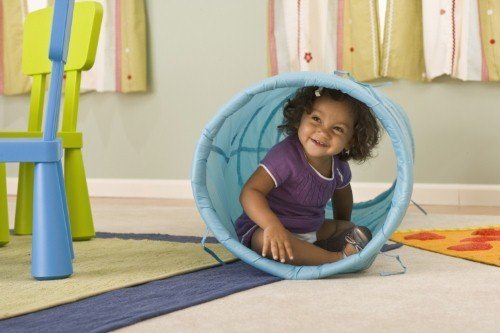 A toddler sitting inside a fabric play tunnel in a comfortable, climate-controlled playroom.
