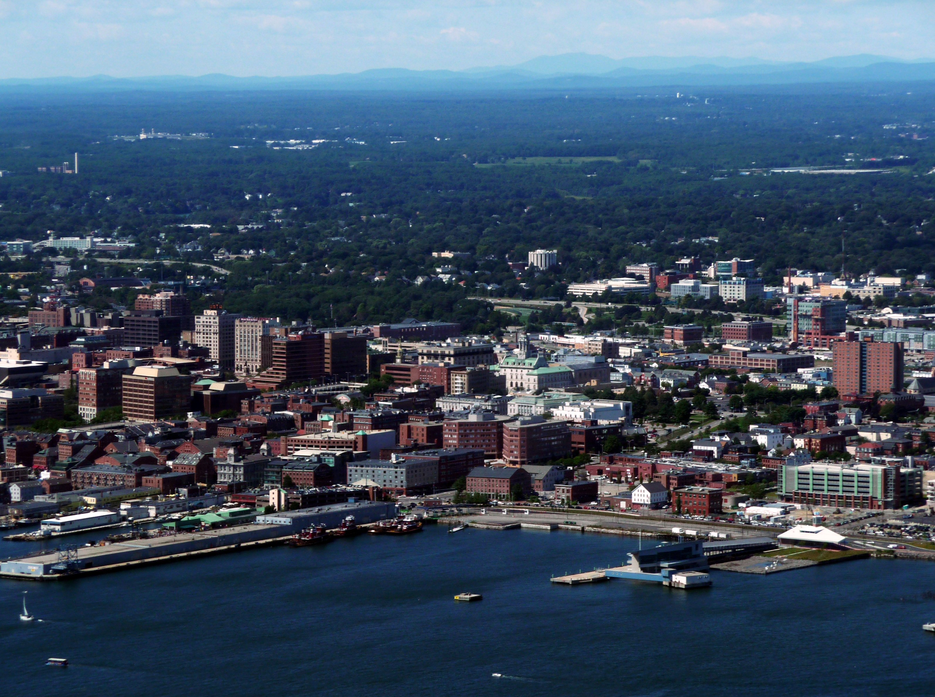 Aerial view of the Portland, Maine skyline featuring the coastal harbor, dense urban buildings, and surrounding suburban landscape.