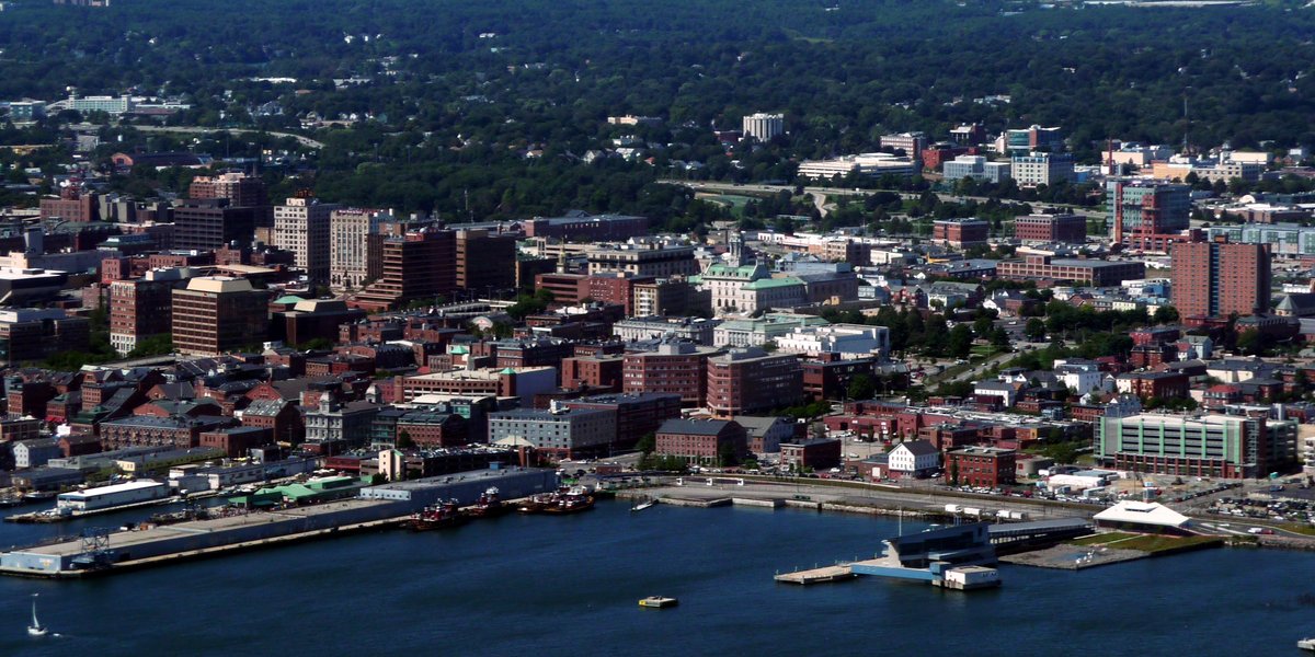 Aerial view of the Portland, Maine skyline featuring the coastal harbor, dense urban buildings, and surrounding suburban landscape.