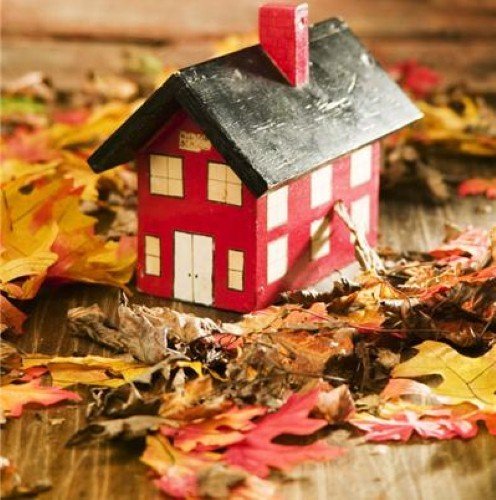 A miniature painted wooden toy house with a red exterior and dark roof displayed among scattered autumn leaves on a rustic wooden surface.