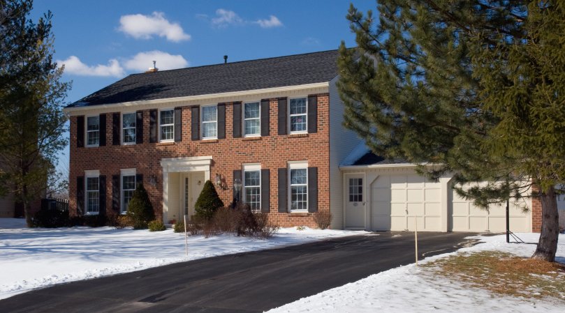 A clear, dry asphalt driveway in front of a snow-covered brick house, demonstrating a radiant heated driveway snow melting system.