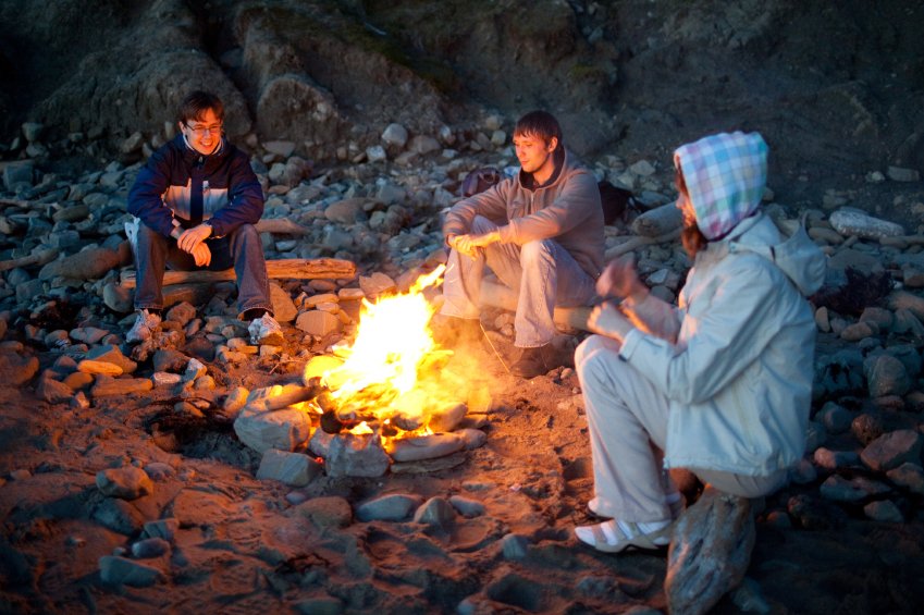 Three people sitting around a campfire on a rocky beach at dusk, illustrating the comfort of warmth in a natural outdoor setting.