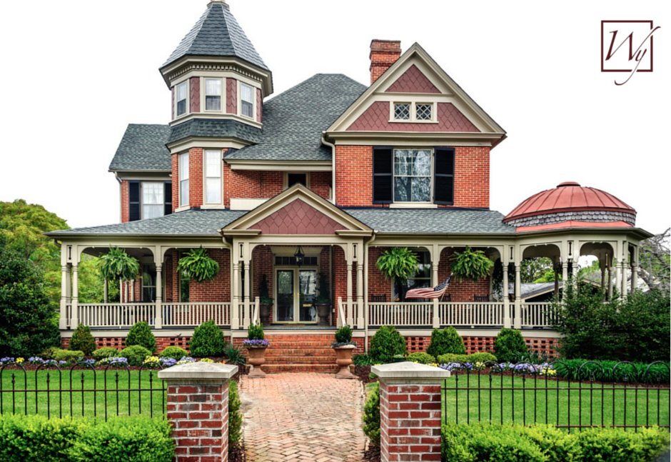 A two-story red brick Queen Anne Victorian home with a turret and wraparound porch, featuring a brick pathway leading to the front entrance.