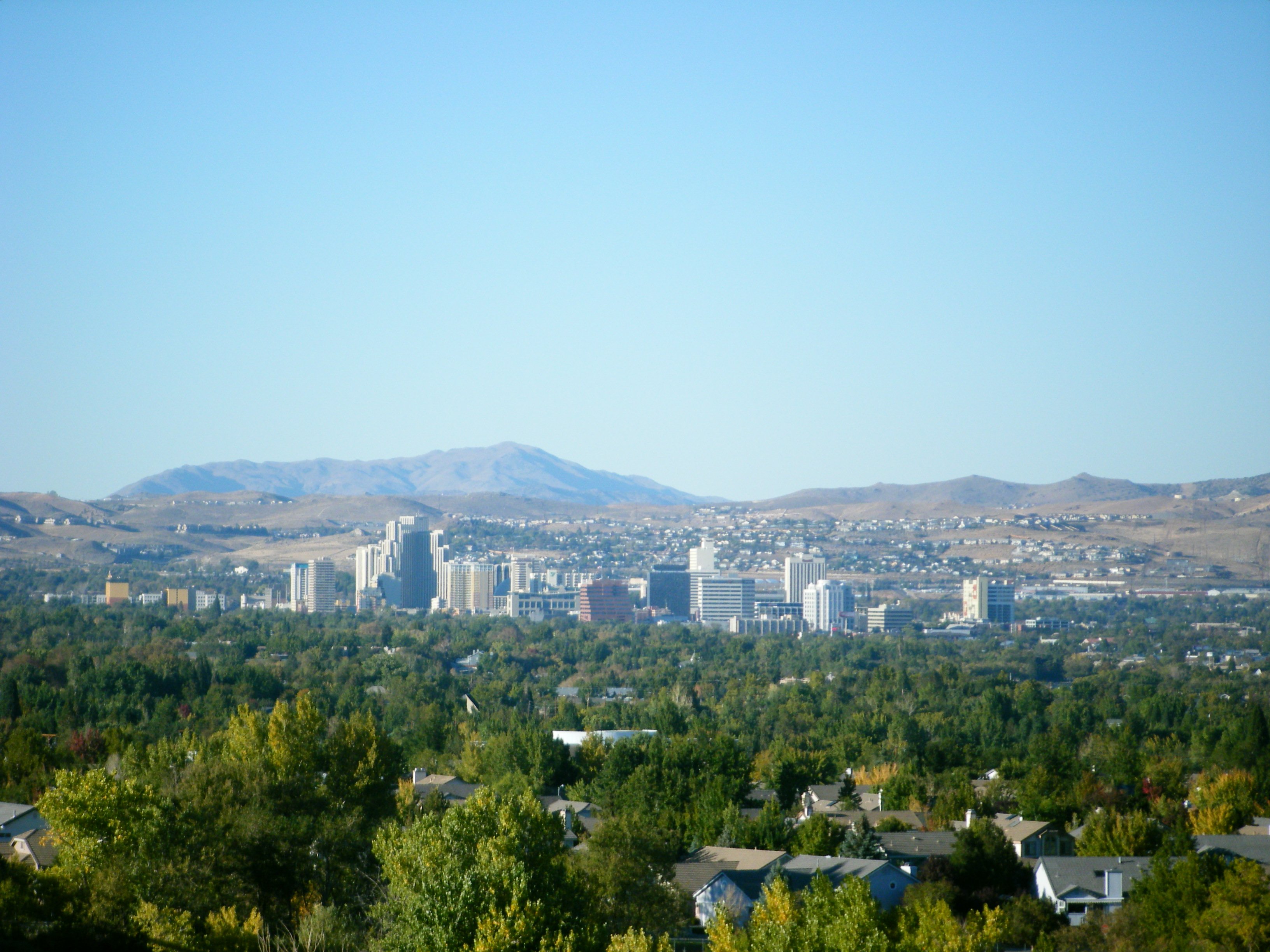 A wide daytime view of the Reno, Nevada cityscape featuring residential rooftops, downtown high-rise buildings, and distant mountain foothills.