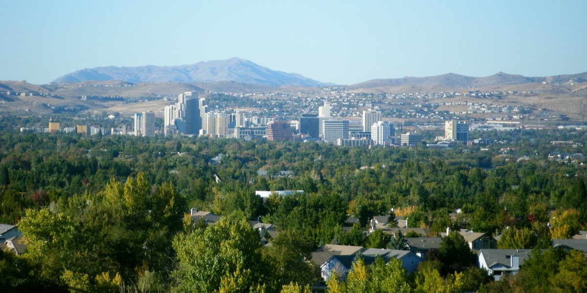 A wide daytime view of the Reno, Nevada cityscape featuring residential rooftops, downtown high-rise buildings, and distant mountain foothills.