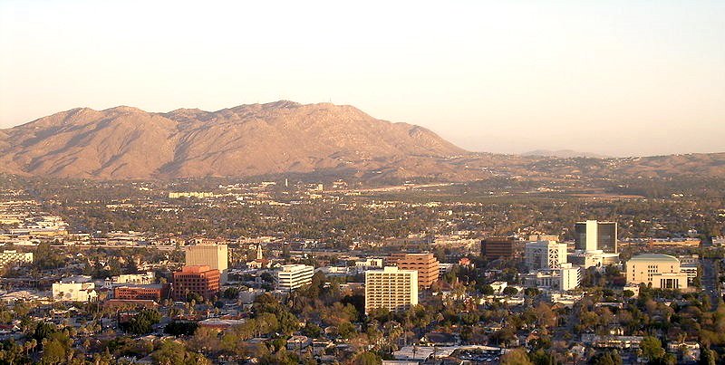 Panoramic view of the Riverside, California skyline featuring mid-rise buildings and urban greenery set against a backdrop of arid mountains.