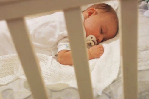 A baby sleeping peacefully on white bedding inside a nursery crib viewed through white wooden slats.