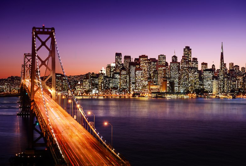 The San Francisco skyline and Bay Bridge illuminated at night with traffic light trails crossing the suspension span under a dusk sky.