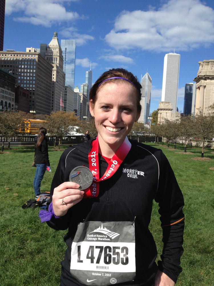 A smiling marathon finisher wearing a race bib and medal stands in a park with the Chicago skyline in the background.