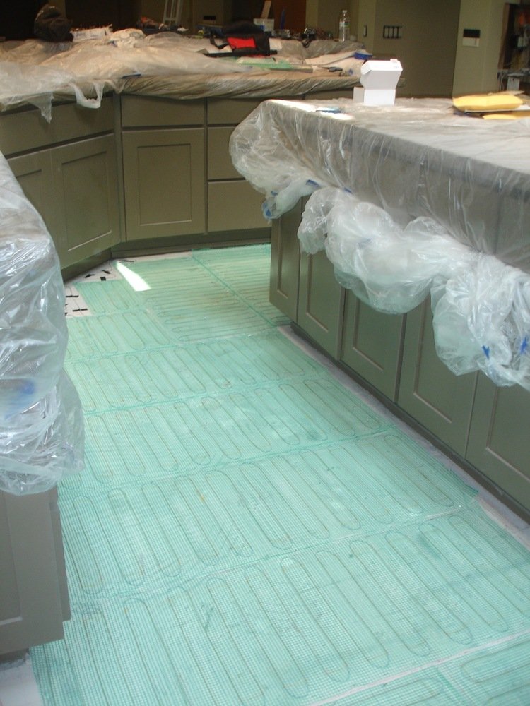TempZone electric radiant floor heating mesh with blue heating cable on green fiberglass mat installed on a kitchen subfloor during a remodel.