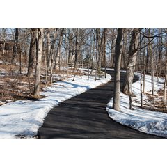 A snow-free asphalt driveway winding through a wooded landscape after a winter storm, maintained by a snow melting system.
