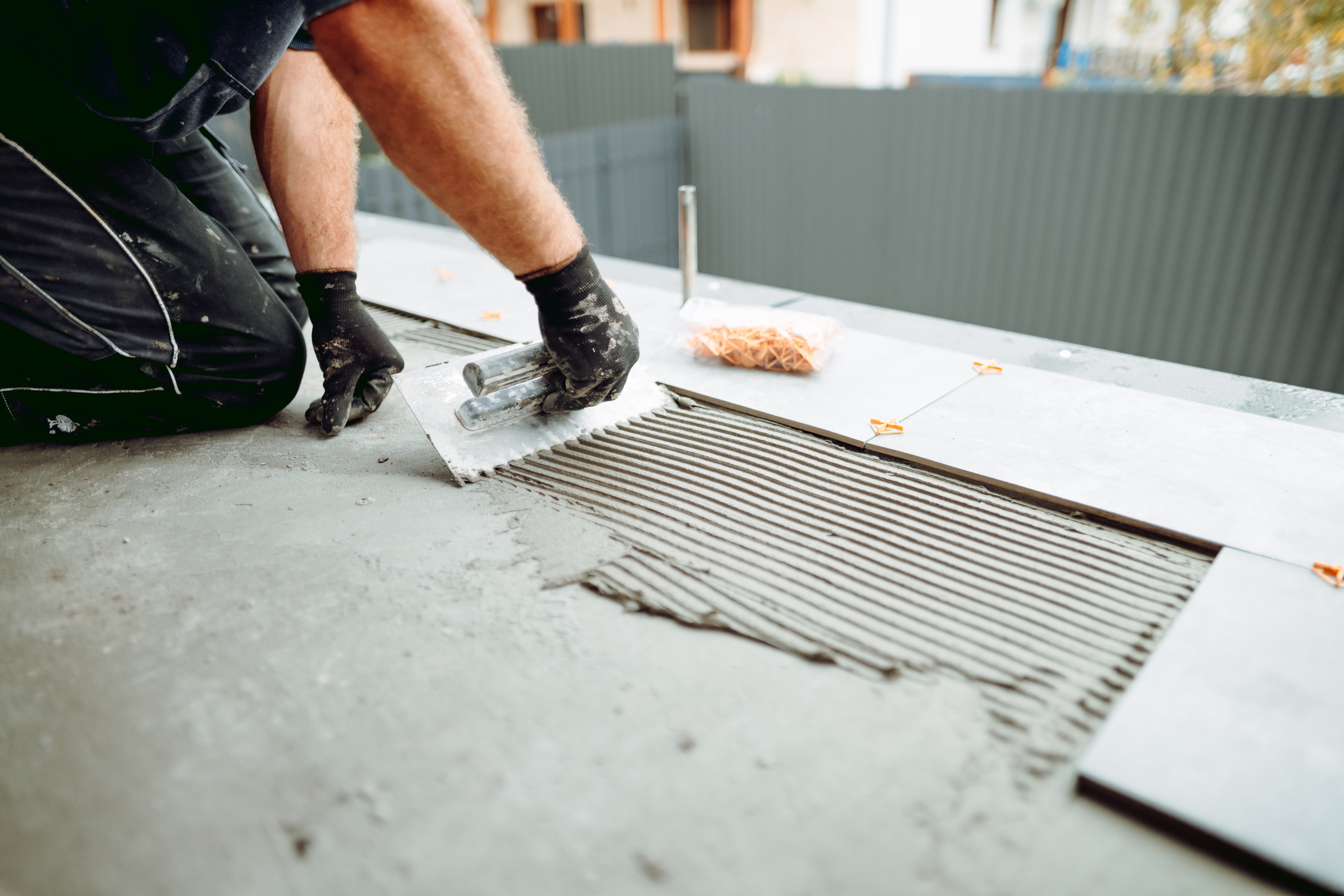 A worker spreading thinset mortar with a notched trowel over a concrete substrate during an outdoor tile installation with leveling spacers.