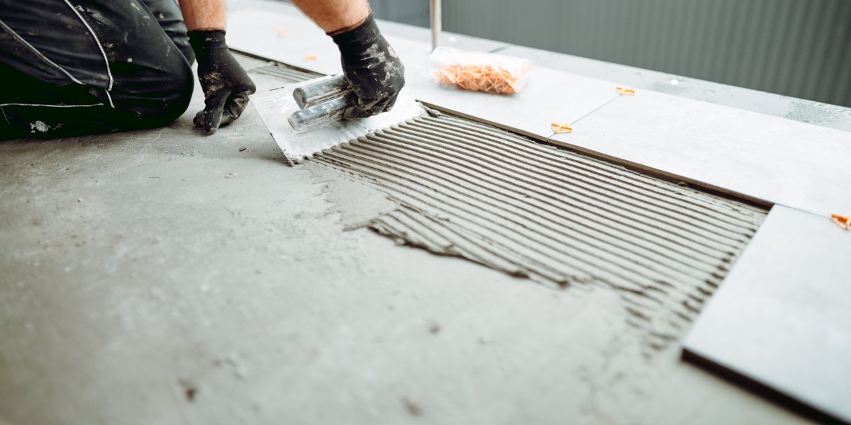 A worker spreading thinset mortar with a notched trowel over a concrete substrate during an outdoor tile installation with leveling spacers.