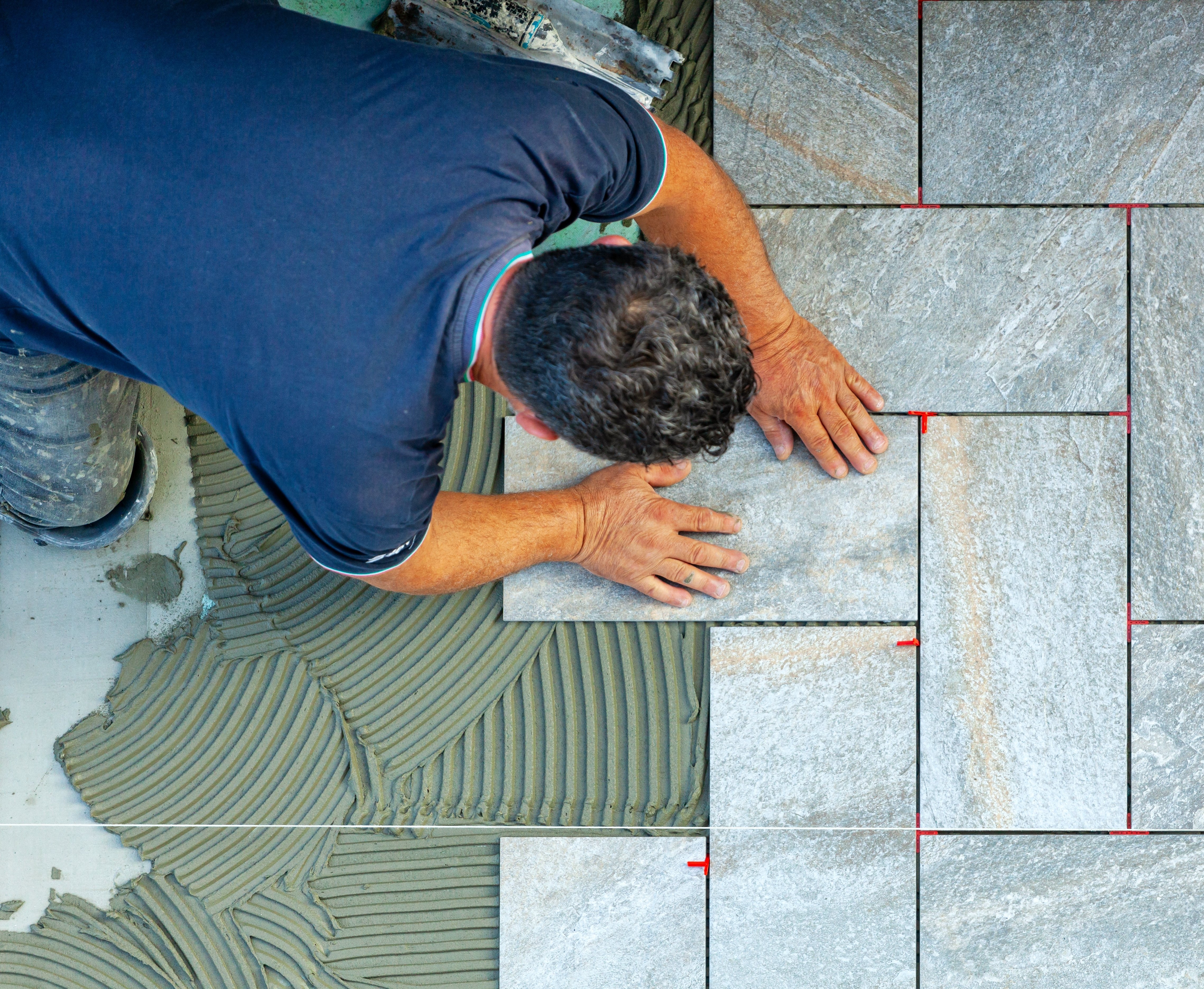 A tile installer sets a grey stone-look tile into a combed bed of wet mortar using red T-shaped spacers for consistent grout lines.