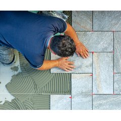 A tile installer sets a grey stone-look tile into a combed bed of wet mortar using red T-shaped spacers for consistent grout lines.