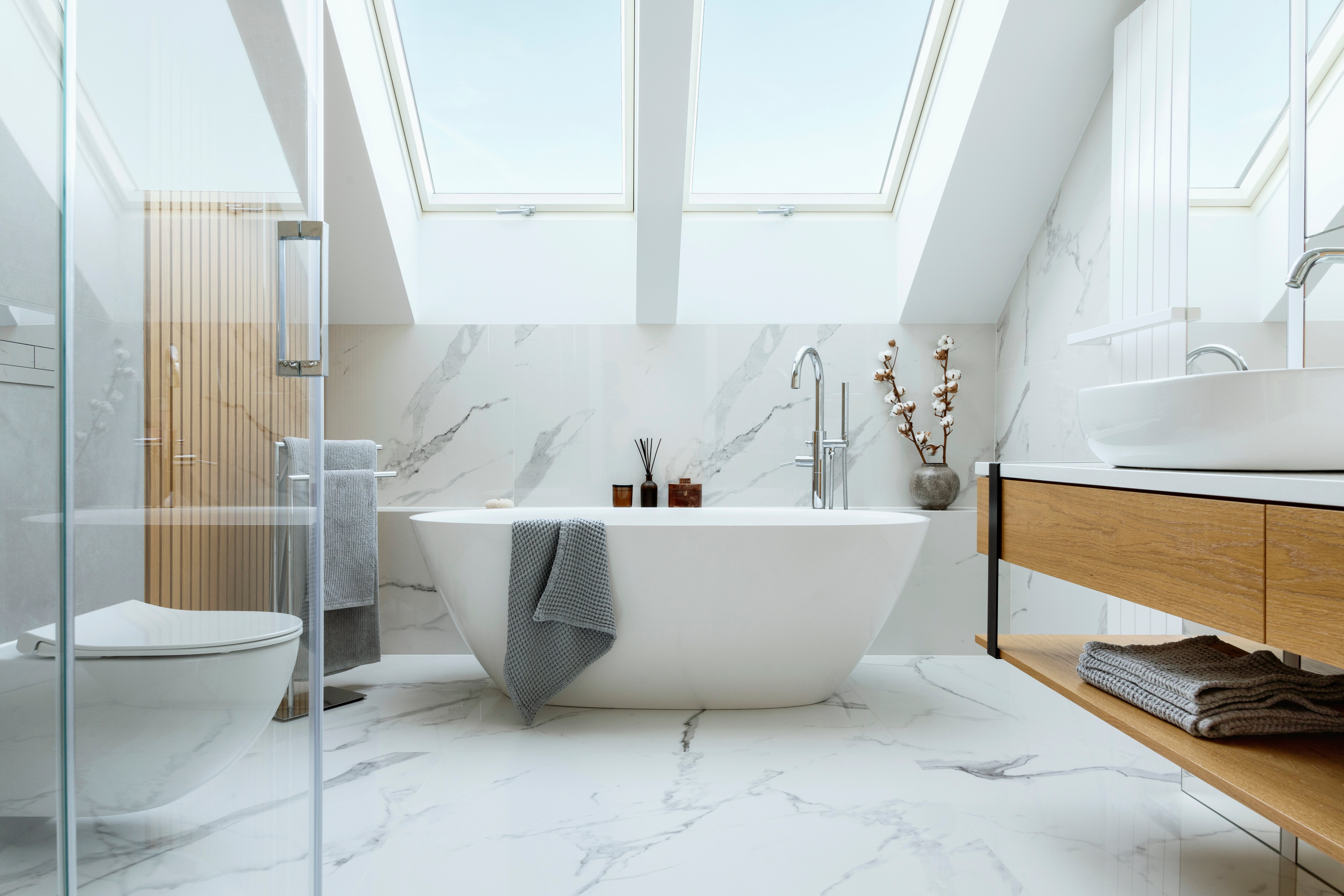 A modern attic bathroom featuring white marble-look tile floors equipped with electric radiant floor heating beneath a freestanding soaking tub.