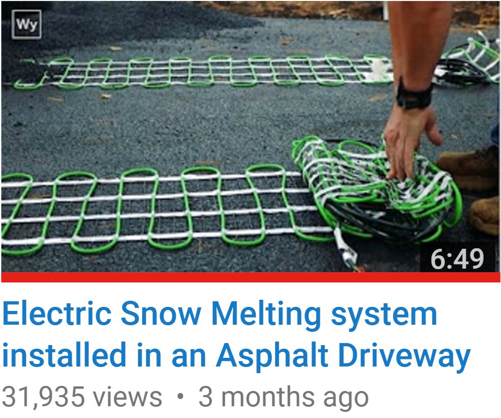 An installer unrolls an electric snow melting mat with green heating cable in a serpentine pattern across an asphalt driveway base.
