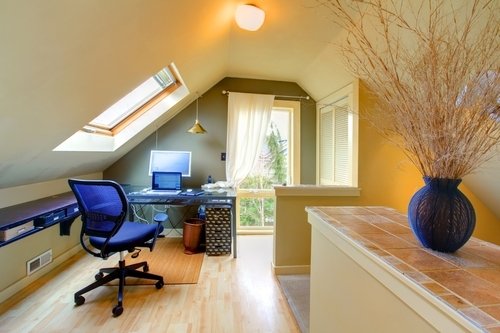 A home office loft with sloped ceilings and light-colored hardwood flooring featuring a desk setup and a terracotta-tiled half-wall.