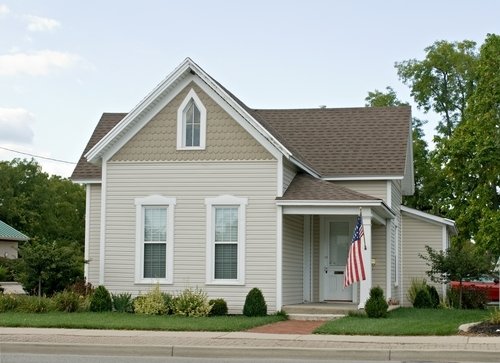 A single-story house with beige lap siding and a gabled roof featuring a front porch and landscaped lawn during a spring improvement season.