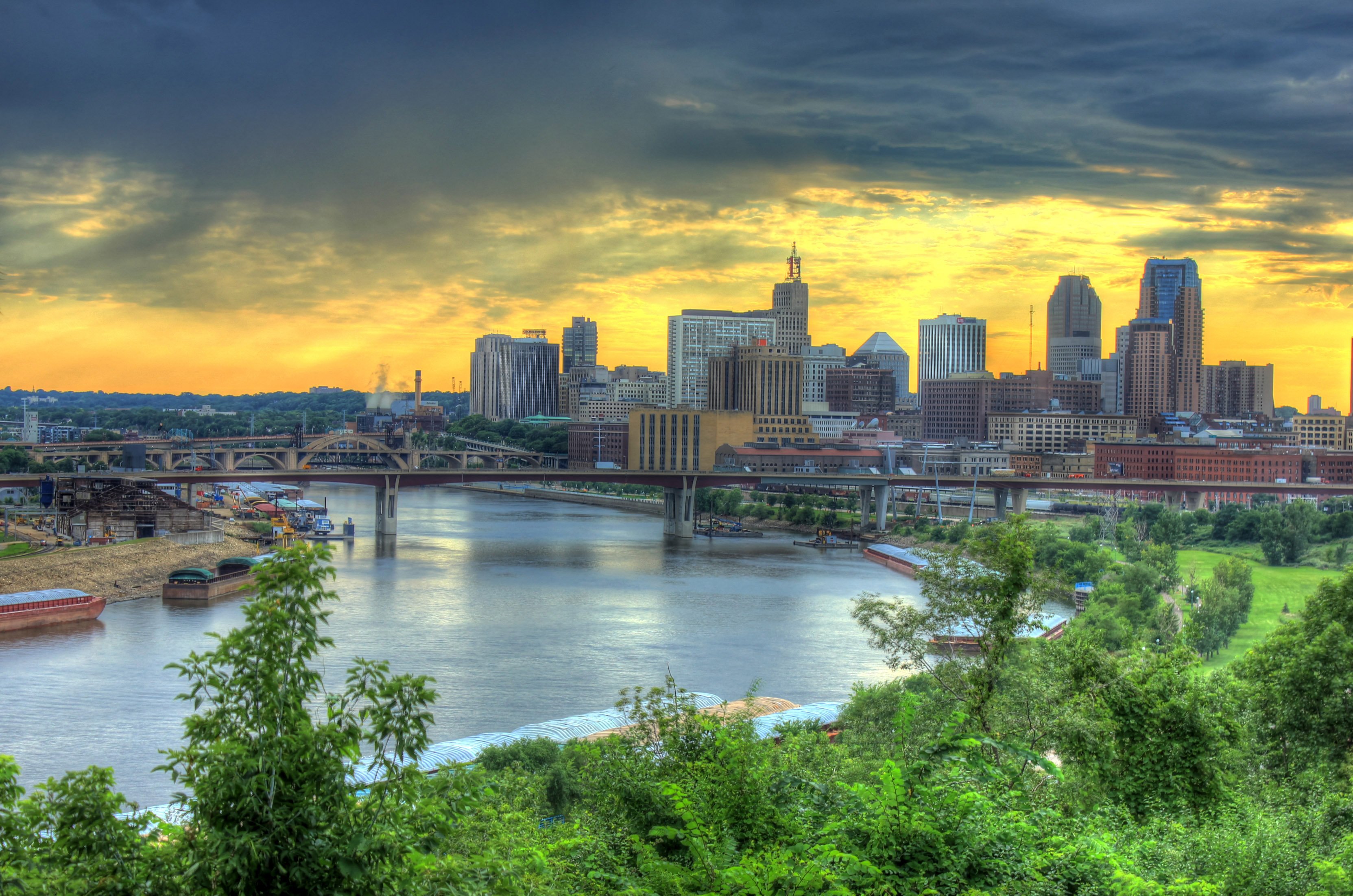 The Saint Paul, Minnesota skyline at sunset with the Mississippi River, bridges, and downtown skyscrapers under a dramatic storm-filled sky.