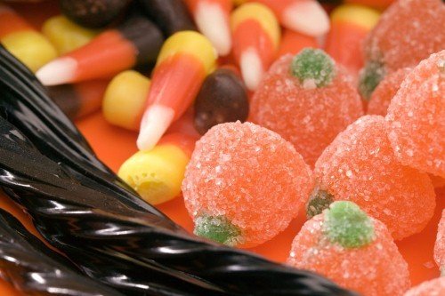 An assortment of Halloween candy including sugar-coated gummy pumpkins, candy corn, and black licorice twists on an orange background.