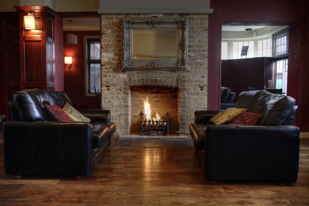 A cozy living room featuring an exposed brick fireplace with a burning fire, framed by two leather sofas and hardwood flooring.