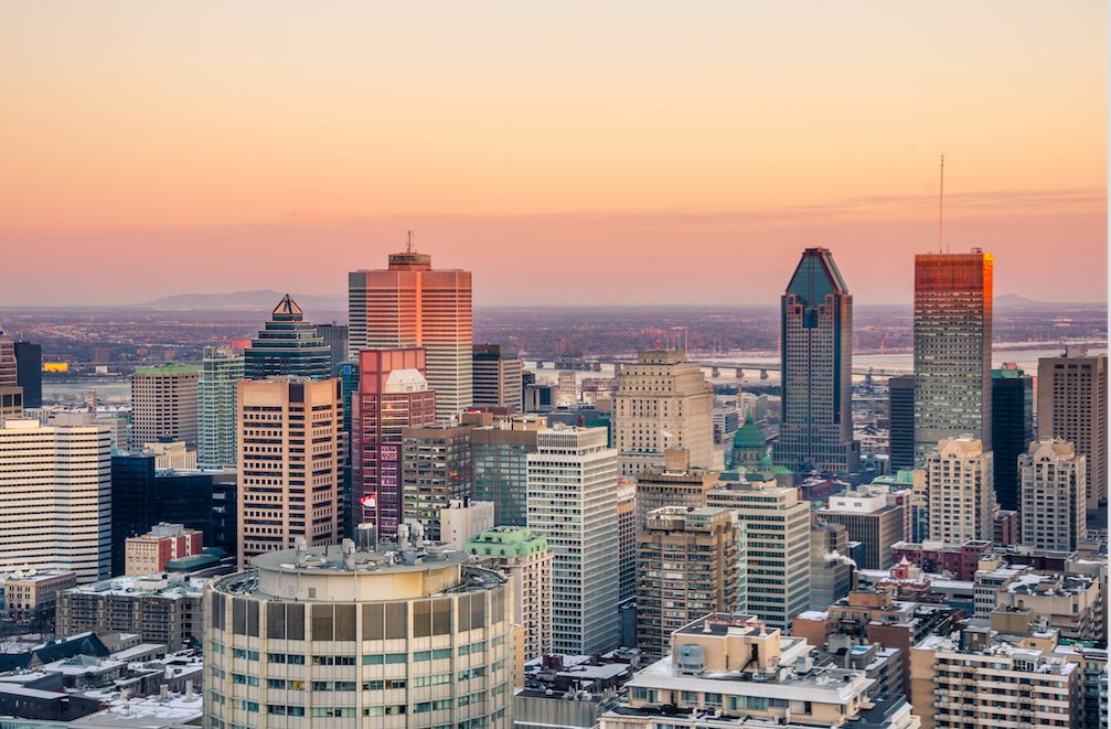 A winter sunset over the Montreal skyline with snow-covered rooftops and illuminated high-rise buildings.