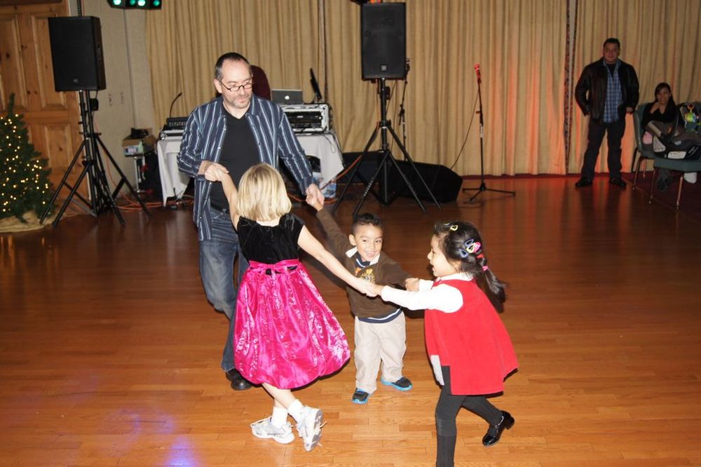 A family dancing on a polished wood floor in a room prepared for a holiday event with a Christmas tree and stage equipment.