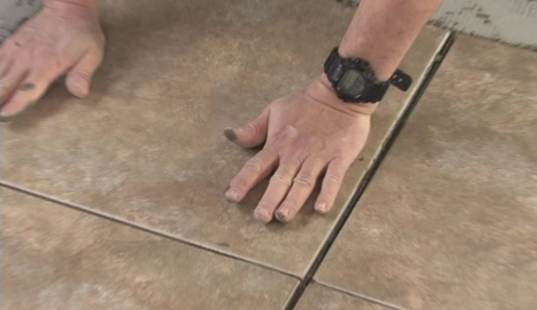 Hands pressing large square beige ceramic floor tiles into thinset mortar during a residential tile installation.