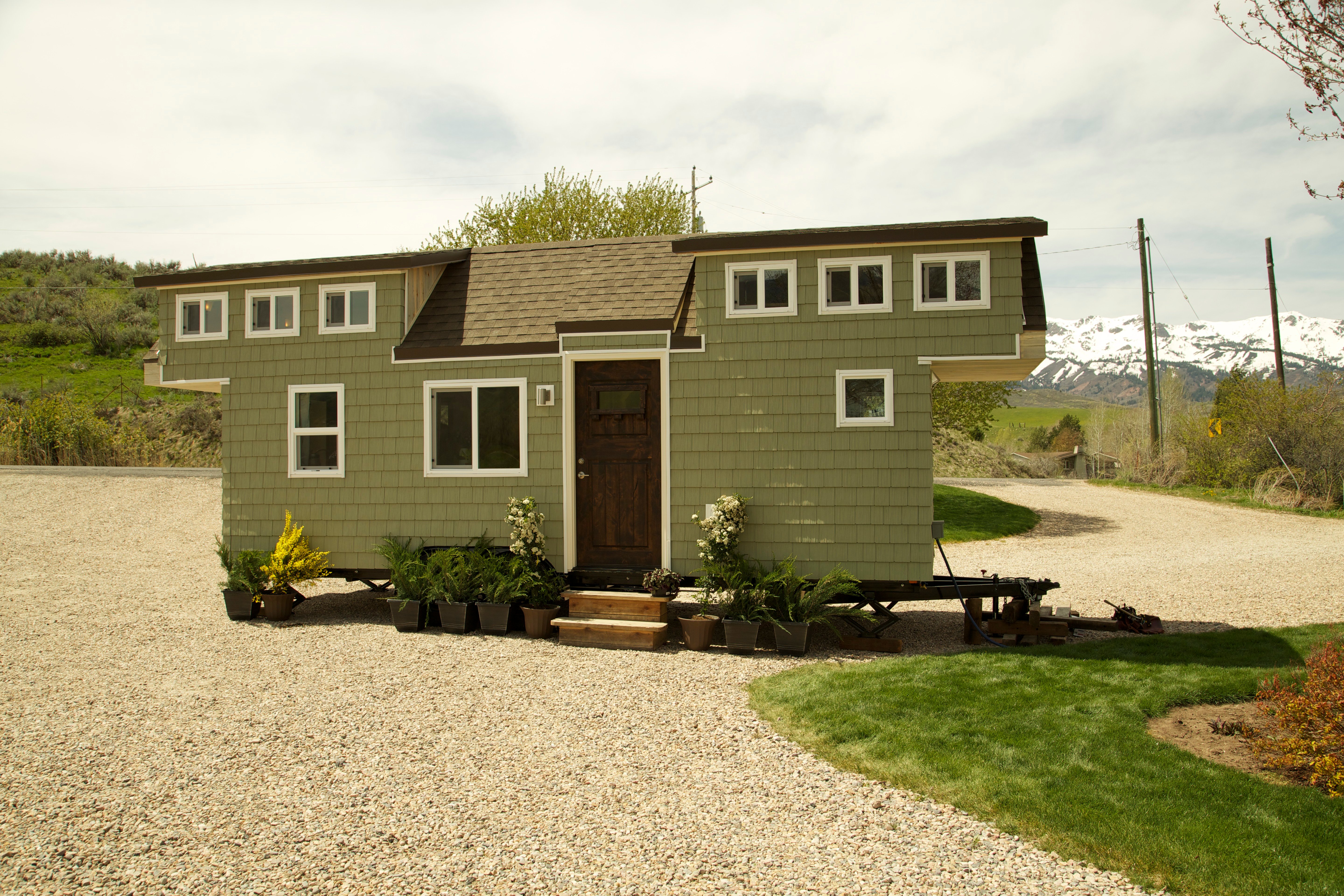 A mobile tiny house with sage-green siding parked on a gravel driveway in front of a mountain landscape.
