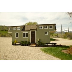 A mobile tiny house with sage-green siding parked on a gravel driveway in front of a mountain landscape.