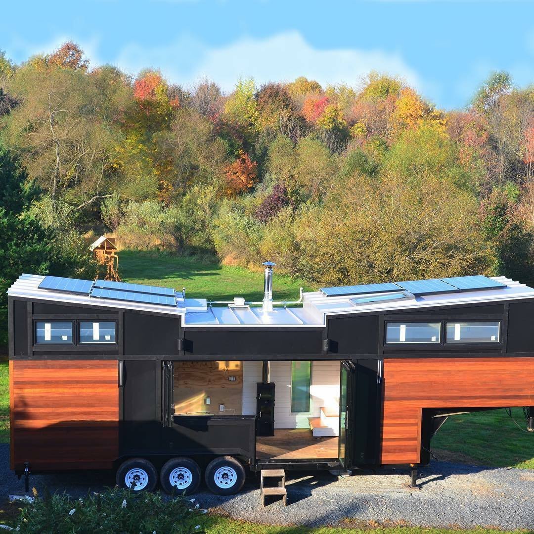 A modern tiny house on wheels with horizontal wood siding and folding glass doors parked in a rural autumn landscape.