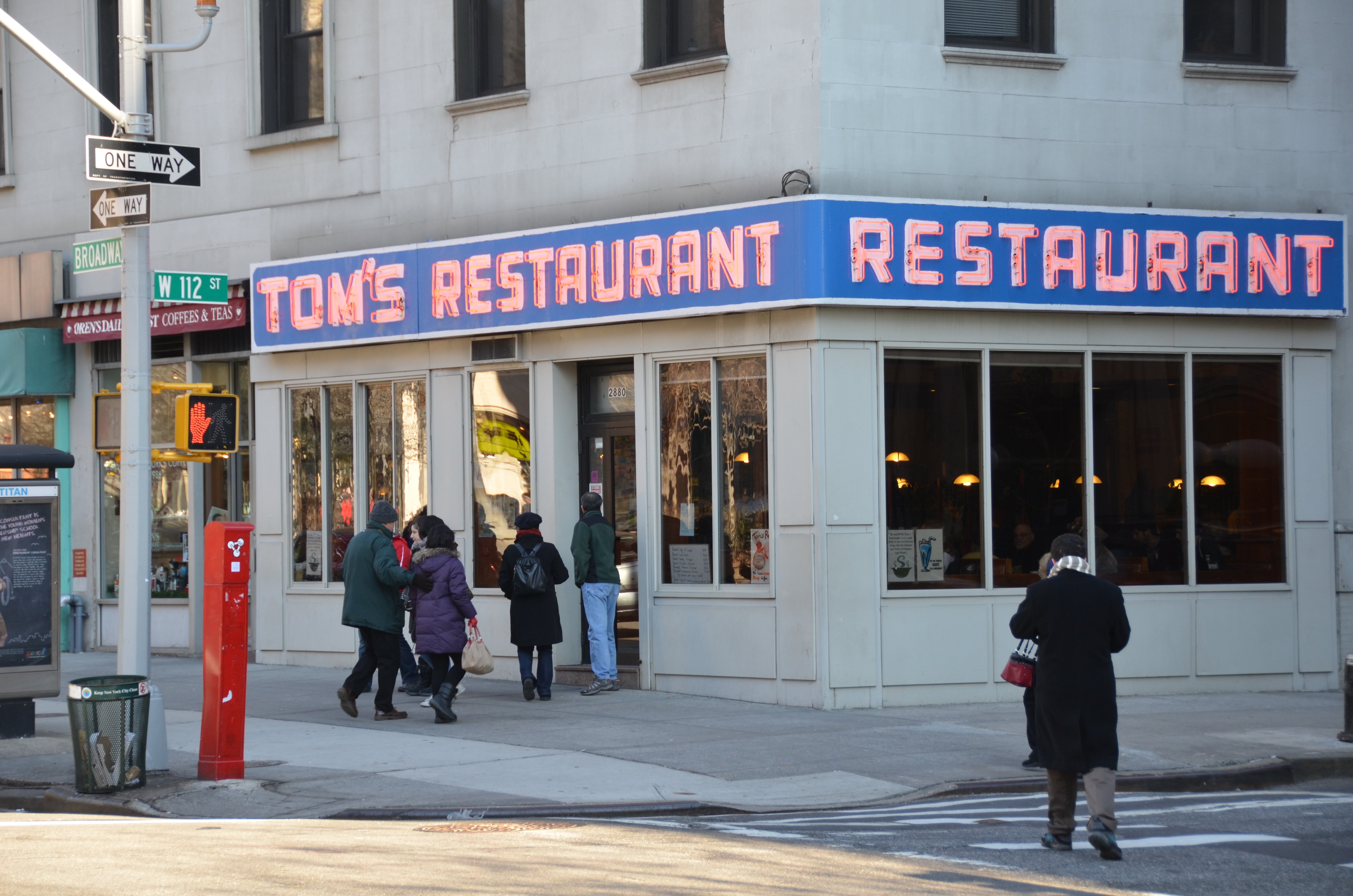 The exterior of Tom's Restaurant on a New York City street corner, featuring its iconic blue fascia sign and large glass windows.