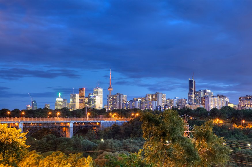 The Toronto skyline at dusk featuring the CN Tower and high-rise buildings behind a steel arch bridge and dense tree foliage.
