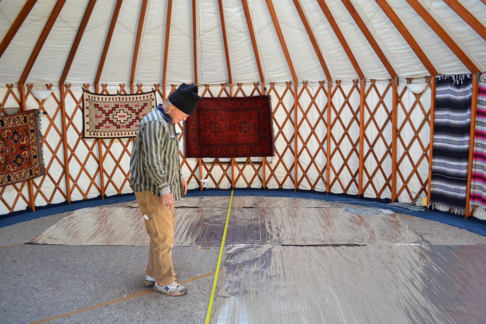 Installer measuring silver foil electric radiant floor heating mats laid over underlayment inside a circular yurt studio.