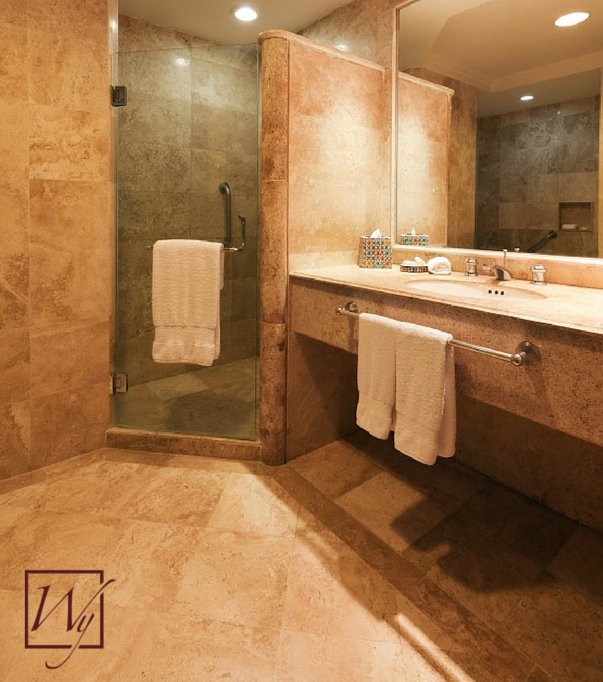 A modern bathroom with beige travertine stone tile flooring and walls, featuring a floating vanity and a glass-door shower enclosure.