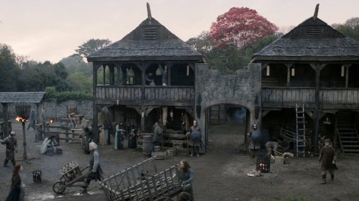 A wide shot of the Winterfell castle courtyard from Game of Thrones featuring stone structures, an arched gateway, and a red-leafed weirwood tree.
