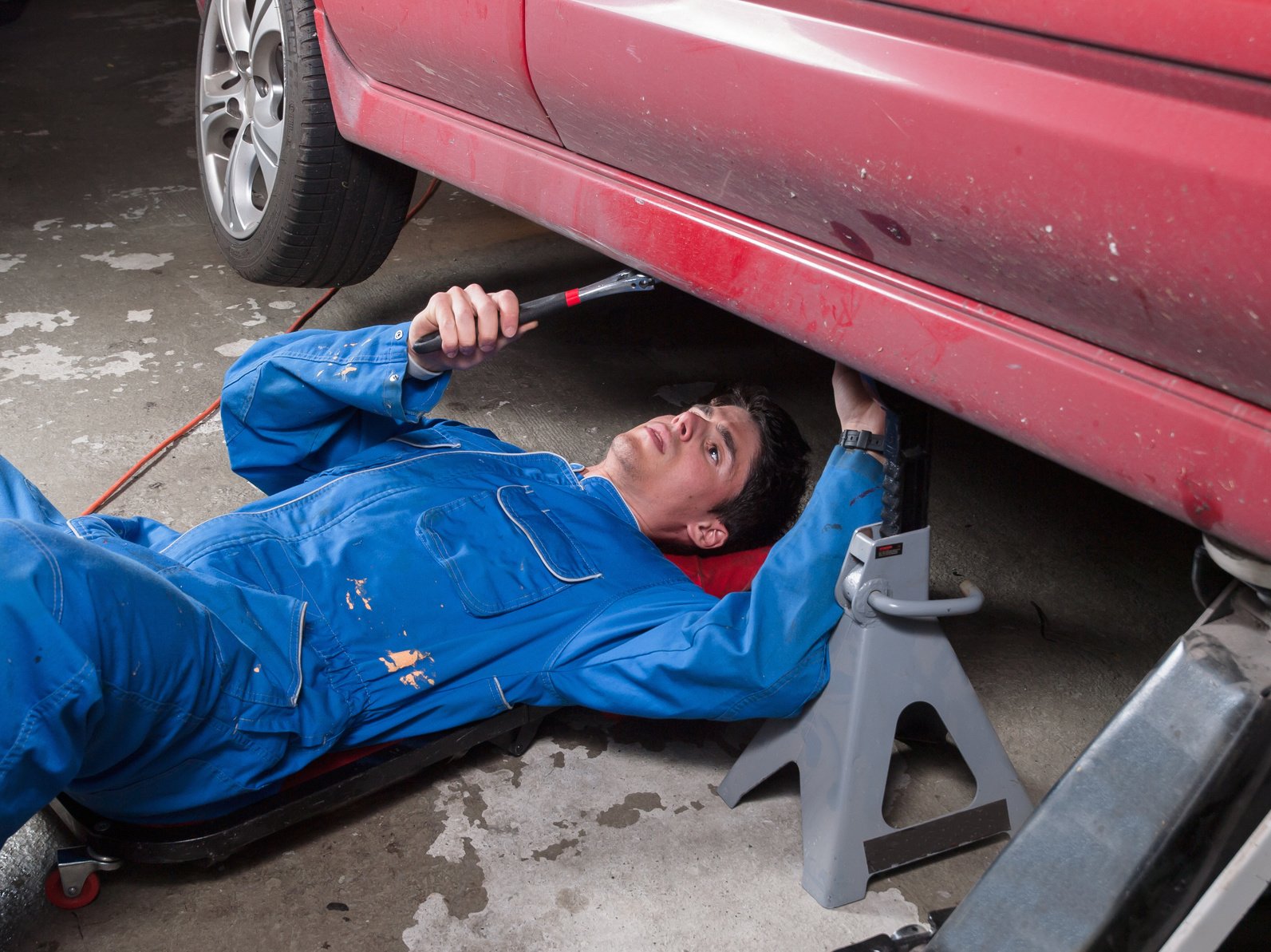 A mechanic in blue overalls uses a tool while lying on a creeper beneath a red car supported by a jack stand on a concrete garage floor.